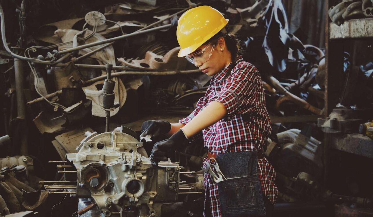 woman wears yellow hard hat holding vehicle part