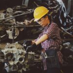 woman wears yellow hard hat holding vehicle part